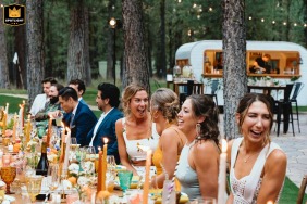 In South Lake Tahoe, California, the wedding party sits at the head table outdoors, laughing together beneath tall trees, with a vintage trailer serving as a drink vendor parked in the background, creating a relaxed and cheerful celebration atmosphere.